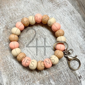 Puede incluir: Un llavero de silicona con cuentas de color rosa y beige con un anillo de llave plateado. Las cuentas tienen un patrón de veta de madera. El llavero está hecho por "Maddy Designs".