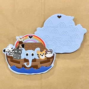 Könnte beinhalten: Ein blauer Silikon-Beißring in Form der Arche Noah mit einem Regenbogen und Tieren darin. Die Rückseite des Beißrings ist blau mit dem Text "Genesis 9:11-17" und "Baby Bundle's Joy Medley".