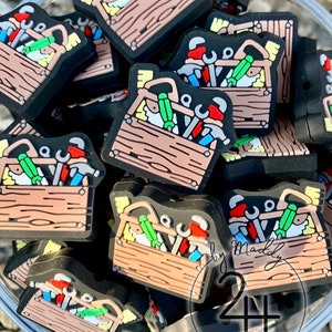 May include: A close-up of a bowl filled with small, colorful, wooden toolbox charms. Each charm features a different assortment of tools, including hammers, screwdrivers, and wrenches. The charms are perfect for adding a touch of whimsy to any project.