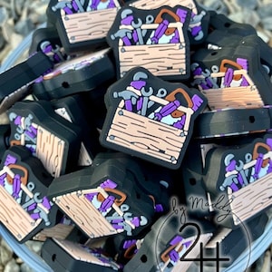 Puede incluir: Primer plano de un mont&oacute;n de dijes de caja de herramientas de color morado y negro. Las cajas de herramientas est&aacute;n hechas de silicona y tienen una variedad de herramientas en su interior, incluyendo un martillo, una llave inglesa y un destornillador. Los dijes est&aacute;n etiquetados como "by Maddy 24+ DESIGNS".
