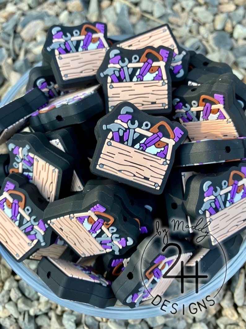 Puede incluir: Primer plano de un mont&oacute;n de dijes de caja de herramientas de color morado y negro. Las cajas de herramientas est&aacute;n hechas de silicona y tienen una variedad de herramientas en su interior, incluyendo un martillo, una llave inglesa y un destornillador. Los dijes est&aacute;n etiquetados como "by Maddy 24+ DESIGNS".