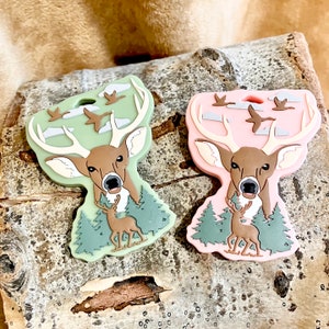 May include: Two silicone deer keychains with a forest scene. One keychain is green and the other is pink. Both keychains feature a deer with antlers, a smaller deer in the background, and flying birds.