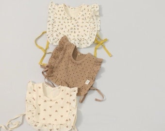 Può includere: Tre bavaglini per neonati con bordi a balze. I bavaglini sono realizzati in un morbido tessuto bianco con diversi motivi: floreale, a pois e a cuori. Ogni bavaglino ha due lunghi lacci per l'allacciatura.