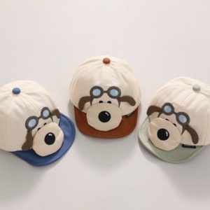 May include: Three beige baseball caps with a cartoon dog face design. Each cap has a different colored brim: blue, brown, and green.