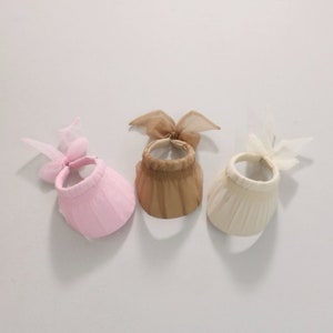 Puede incluir: Tres viseras solares con lazos. Las viseras son de color rosa, beige y marrón. Cada visera tiene un lazo a juego en la parte superior.