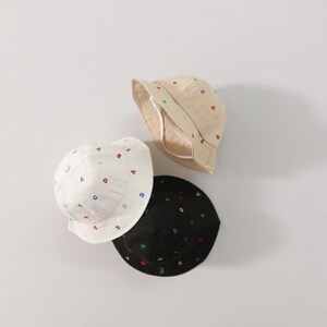 May include: Three bucket hats in beige, white, and black. Each hat is embroidered with colorful letters. The hats are made of a soft fabric and have a wide brim. The hats are designed for children.