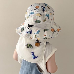 May include: White sun hat with a dinosaur pattern and a matching bib. The hat features a wide brim and a drawstring. The bib has three dinosaur patches. The hat and bib are designed for children.