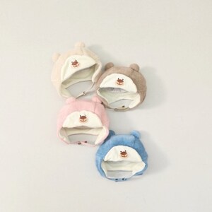 May include: Four knitted baby hats in beige, brown, pink, and blue, each with a bear face design. The hats have ear flaps and a button closure under the chin. The hats are designed for warmth and comfort.