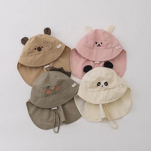May include: Four children's sun hats in tan, pink, olive green, and cream. Each hat features a different animal design, including a bear, panda, dinosaur, and a smiling face. The hats have a wide brim and neck flap for sun protection.