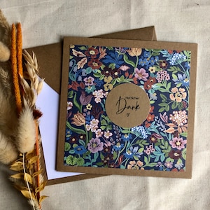 Könnte beinhalten: Eine florale Grußkarte mit marineblauem Hintergrund und einer Vielzahl bunter Blumen. Die Karte hat einen braunen Rand und einen runden Aufkleber mit den Worten "Herzlichen Dank" und einem Herz. Ein Umschlag und eine leere weiße Karte sind ebenfalls sichtbar.