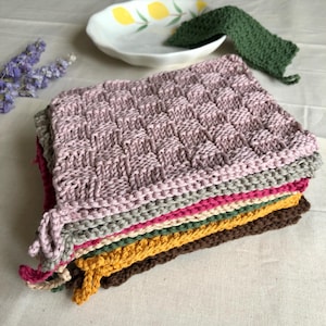 Pode incluir: Uma pilha de panos de lavagem coloridos e tecidos em tons de rosa, cinza, amarelo e marrom. O pano superior tem um padrão texturizado de tecido de cesta. Um pano de lavagem verde e um prato branco com um desenho de limão estão no fundo.