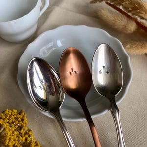 Könnte beinhalten: Drei Löffel mit Baumdesigns liegen auf einem weißen Teller. Die Löffel sind silber- und kupferfarben. Im Hintergrund befindet sich eine weiße Tasse. Die Löffel sind wahrscheinlich zum Servieren oder Essen.