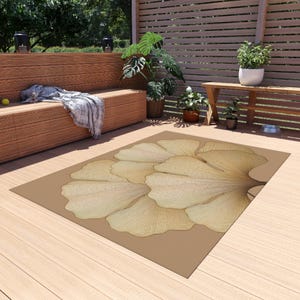 May include: A rectangular outdoor rug featuring a large, stylized ginkgo leaf design in shades of beige and tan. The rug is placed on a wooden deck, with a bench and plants in the background.