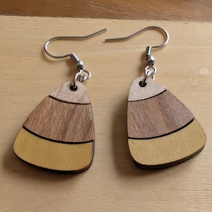 Puede incluir: Un par de pendientes de madera con diseño de caramelo de maíz. Cada pendiente tiene forma triangular con tres franjas horizontales: beige claro, marrón medio y amarillo, separadas por finas líneas negras. Los pendientes tienen ganchos plateados.