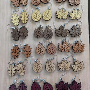 Puede incluir: Un conjunto de pendientes de madera con forma de hojas en varios tonos de marrón y burdeos. Los pendientes presentan diseños de hojas de roble, arce y lágrima. Con ganchos plateados. Sobre una superficie de madera clara.