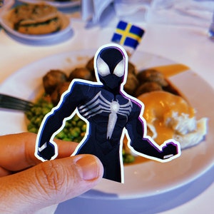 May include: A black and white Spider-Man sticker is held in front of a plate of food. The food includes meatballs, peas, gravy, and mashed potatoes.