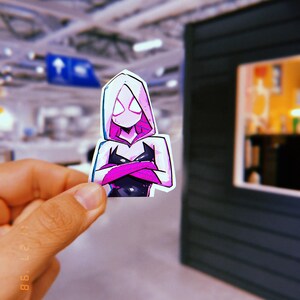 May include: A sticker of a female superhero wearing a pink hooded costume with black and white accents. The superhero has a white mask with black eyes and is standing with her arms crossed.