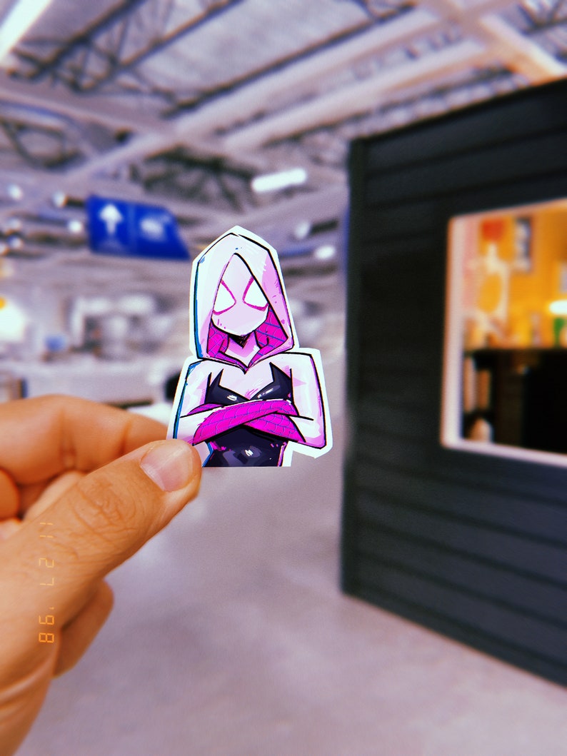 May include: A sticker of a female superhero wearing a pink hooded costume with black and white accents. The superhero has a white mask with black eyes and is standing with her arms crossed.