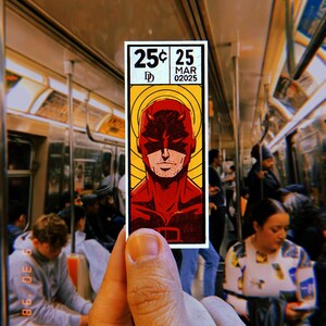 Peut inclure: Un ticket de m&eacute;tro de style bande dessin&eacute;e rouge et jaune avec le texte "25&cent; 25 MAR 02025" et une image de bande dessin&eacute;e de Daredevil, un super-h&eacute;ros en costume rouge et masque rouge.