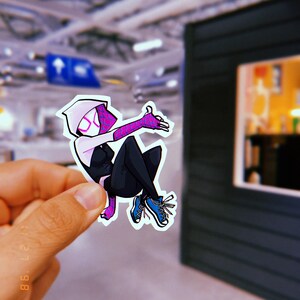 May include: A white and black cartoon sticker of Spider-Gwen, a female superhero, with pink accents, wearing a white hooded suit and blue trainers. She is in a jumping pose with her right arm raised and her left arm extended.
