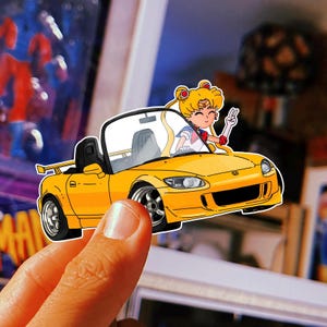 Può includere: Un adesivo fustellato raffigurante un'auto sportiva gialla con un personaggio dei cartoni animati che saluta dal posto di guida. L'auto ha dettagli neri e ruote argentate. Il personaggio ha i capelli biondi e indossa un'uniforme da marinaio.