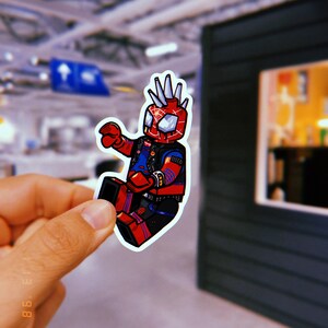 May include: A red and blue cartoon sticker of a superhero with spiky hair, wearing a black leather jacket and holding a web shooter. The sticker is on a white background.
