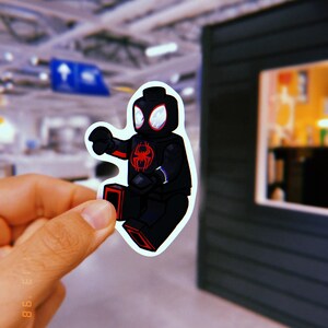 Puede incluir: Una minifigura Lego de Spider-Man negra y roja, vestida con un traje negro con detalles rojos. La minifigura sostiene un lanzatelara&ntilde;as en su mano derecha y tiene un s&iacute;mbolo de ara&ntilde;a rojo en su pecho.