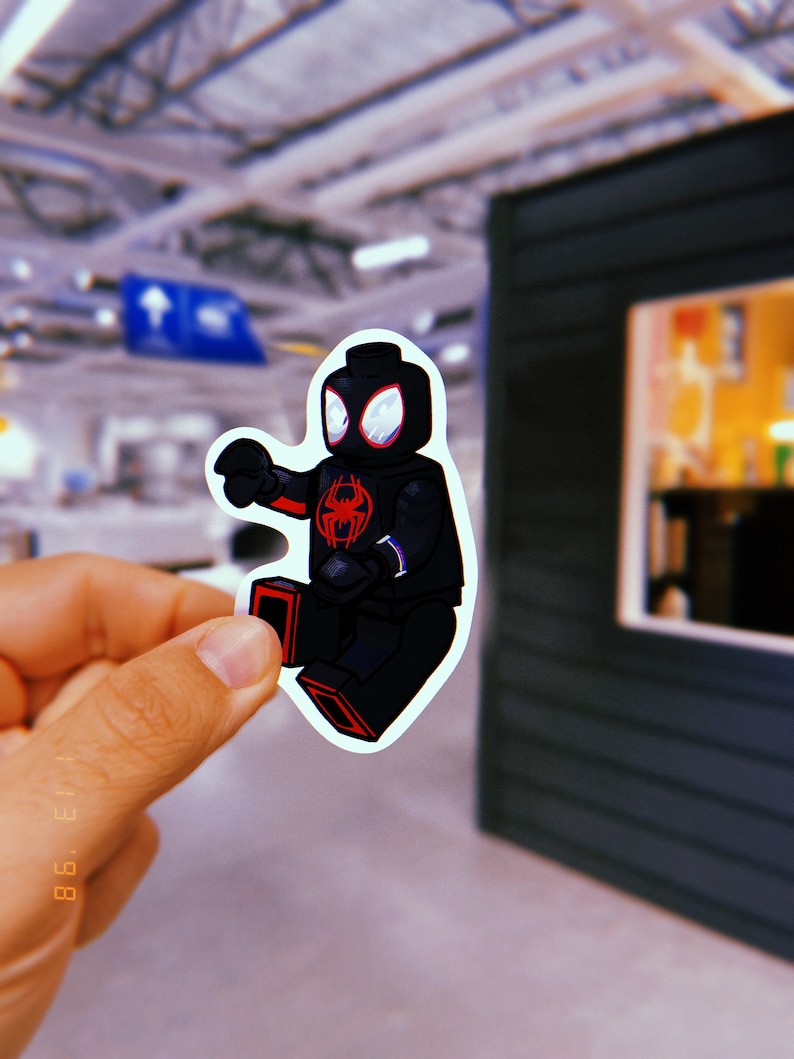 Puede incluir: Una minifigura Lego de Spider-Man negra y roja, vestida con un traje negro con detalles rojos. La minifigura sostiene un lanzatelara&ntilde;as en su mano derecha y tiene un s&iacute;mbolo de ara&ntilde;a rojo en su pecho.