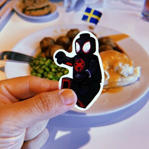 Puede incluir: Una minifigura Lego negra y roja de Spider-Man, sosteniendo un lanzador de telara&ntilde;as rojo en su mano derecha, est&aacute; en un plato blanco con una bandera sueca en el fondo. El plato tiene pur&eacute; de patatas, salsa, guisantes y alb&oacute;ndigas.