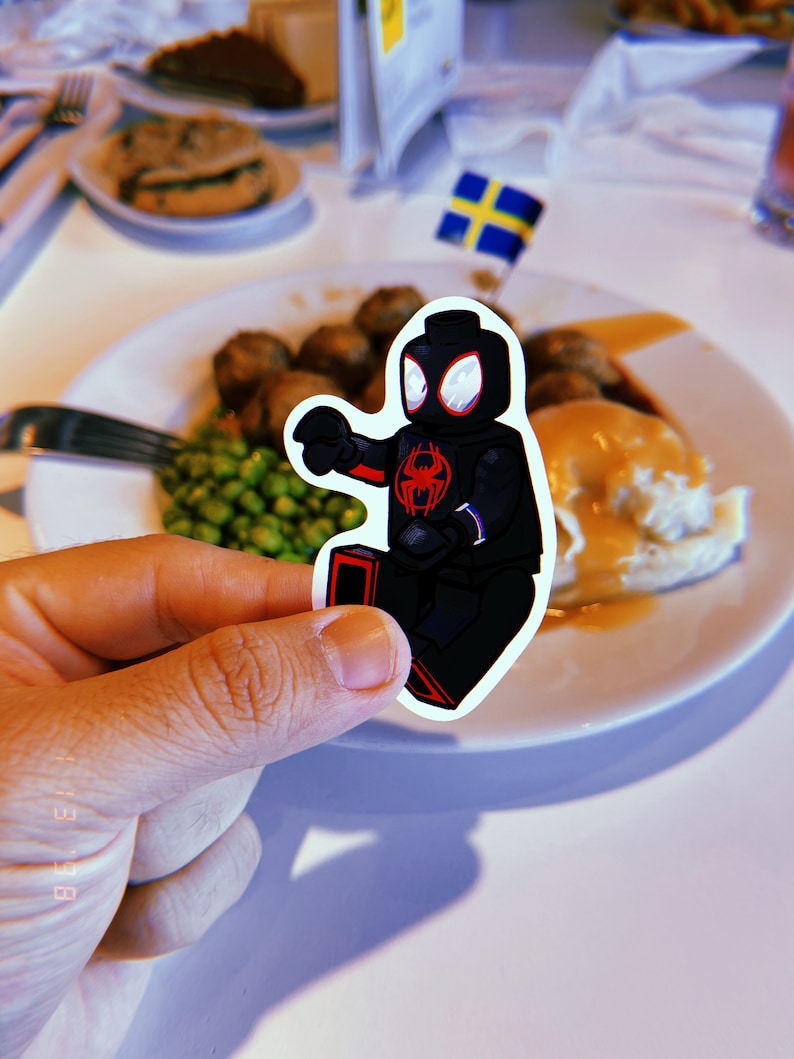 Puede incluir: Una minifigura Lego negra y roja de Spider-Man, sosteniendo un lanzador de telara&ntilde;as rojo en su mano derecha, est&aacute; en un plato blanco con una bandera sueca en el fondo. El plato tiene pur&eacute; de patatas, salsa, guisantes y alb&oacute;ndigas.