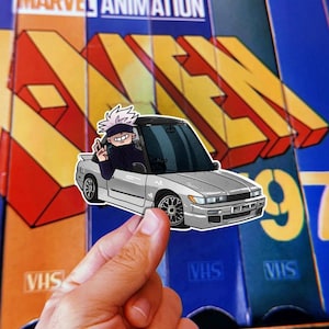 Puede incluir: Una pegatina que representa un coche plateado con un personaje de dibujos animados dentro, saludando. La pegatina se sostiene sobre un fondo de cintas VHS y el texto "MARVEL ANIMATION" y "X-MEN".