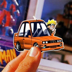 May include: Orange car sticker featuring a cartoon car with a character from an anime series. The car has a sporty design with black windows and a smiling character waving from the driver's side. The sticker is held by a person's hand.