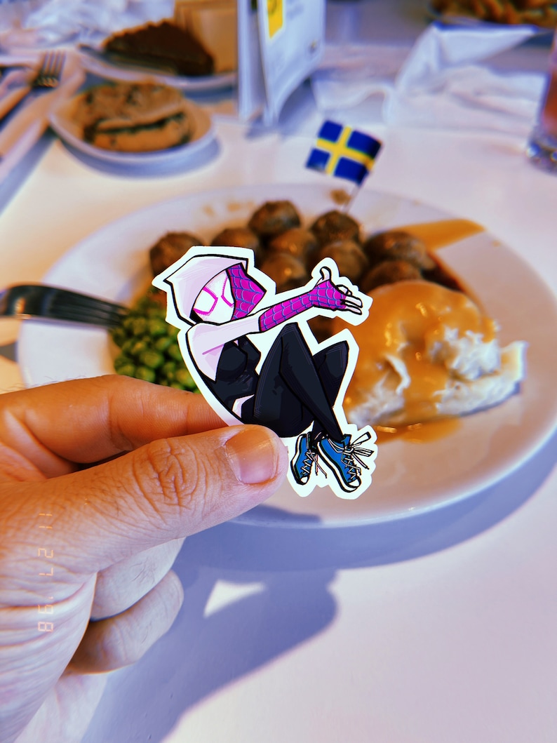 May include: A sticker of a cartoon character wearing a pink and black suit with blue shoes. The character is holding a hand up in a wave. The sticker is being held over a plate of food.