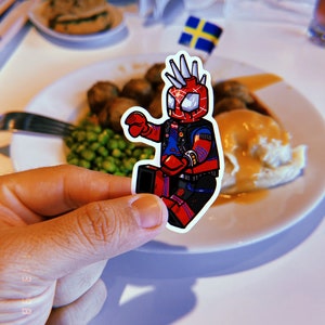 May include: A sticker of a cartoon superhero wearing a red and blue suit with a black leather jacket and a mohawk. The sticker is placed on a white plate with a meal of mashed potatoes, gravy, peas, and meatballs.