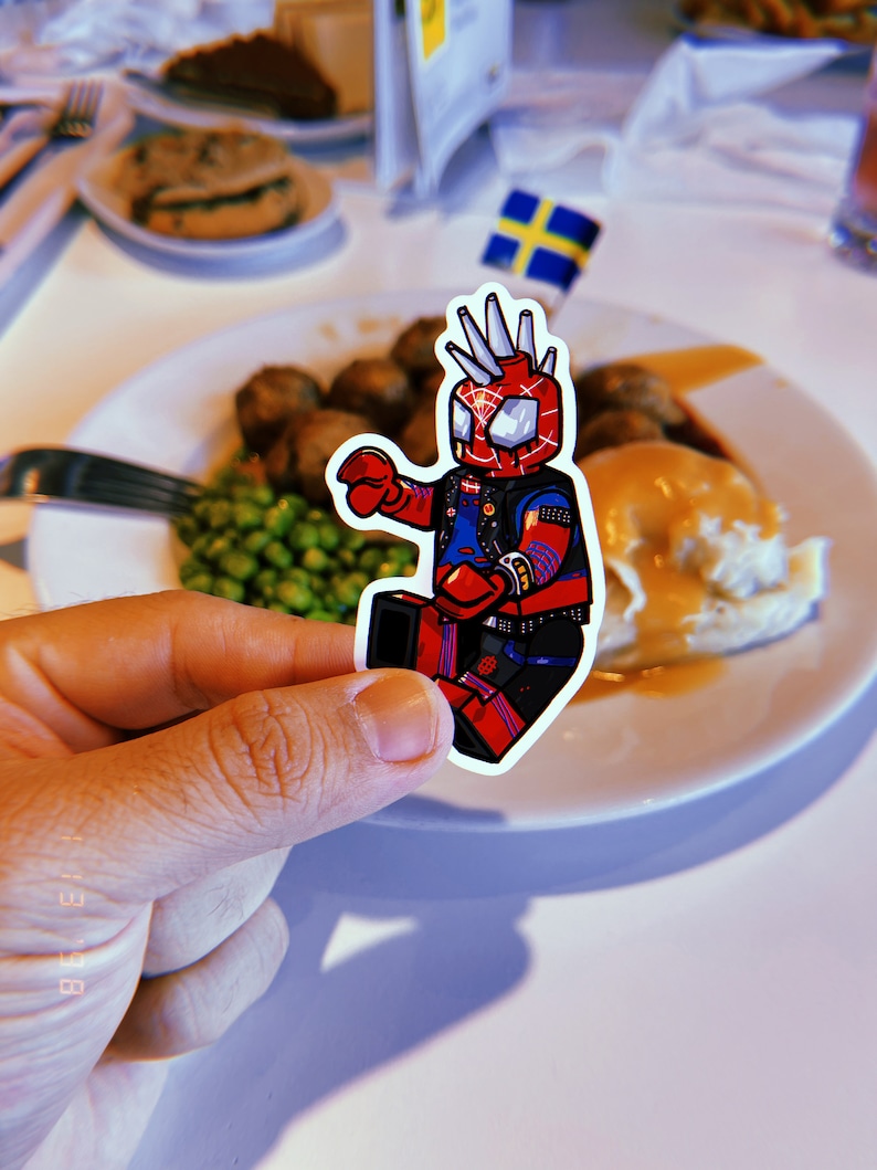 May include: A sticker of a cartoon superhero wearing a red and blue suit with a black leather jacket and a mohawk. The sticker is placed on a white plate with a meal of mashed potatoes, gravy, peas, and meatballs.