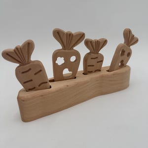 Könnte beinhalten: Holzspielzeug mit vier geschnitzten Karotten in einem Holzsockel. Die Karotten haben detaillierte Blattoberteile und geschnitzte Linien. Eine Karotte hat einen blütenförmigen Ausschnitt, eine andere zwei kreisförmige Ausschnitte. Das Spielzeug ist aus hellem Holz gefertigt.