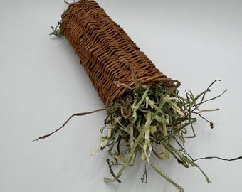 May include: A brown woven wicker tube filled with dried hay. The tube is cylindrical and appears to be hand-woven. The hay is a mix of green and tan, with some strands extending beyond the tube's opening. This item is likely a pet toy.