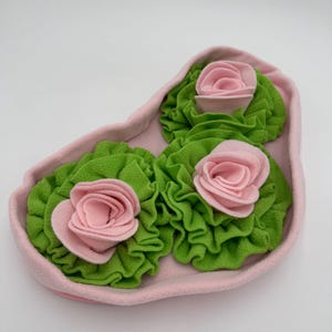 Pode incluir: Uma bandeja de feltro rosa com três rosas de feltro. Cada rosa tem um centro rosa e folhas verdes frisadas. A bandeja é de cor rosa claro e tem uma borda ondulada. As rosas estão uniformemente espaçadas.