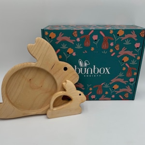 Puede incluir: Dos bandejas de madera con forma de conejito, una grande y otra pequeña, frente a una caja turquesa. La caja está decorada con conejitos, flores y mariposas. La caja tiene el texto "bunbox SOCIETY".