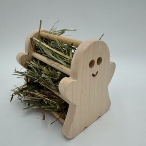 Op de afbeelding: Een houten hooi-voederbak in de vorm van een spook voor kleine dieren. De voederbak heeft een gleuf voor hooi en een uitsnijding van een spookgezicht. De voederbak is gevuld met gedroogd gras.