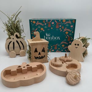 Op de afbeelding: Een houten hooiruif in de vorm van een pompoen, een houten hooiruif in de vorm van een spook, een houten pompoen-puzzel, een houten pompoen-voedselbak en een houten hooiruif in de vorm van een jack-o'-lantern. De items zijn allemaal onderdeel van een Halloween-thema konijnen speelgoedset. De doos op de achtergrond is gelabeld "bunbox society".