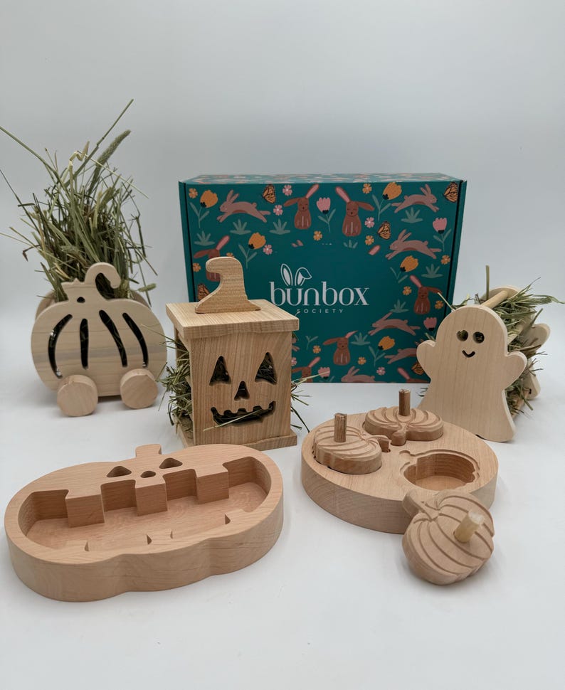 Op de afbeelding: Een houten hooiruif in de vorm van een pompoen, een houten hooiruif in de vorm van een spook, een houten pompoen-puzzel, een houten pompoen-voedselbak en een houten hooiruif in de vorm van een jack-o'-lantern. De items zijn allemaal onderdeel van een Halloween-thema konijnen speelgoedset. De doos op de achtergrond is gelabeld "bunbox society".