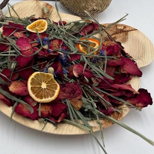 May include: A wooden leaf-shaped bowl filled with dried rose petals, green grass, dried orange slices, and small blue flowers. The bowl is light brown and sits on a white surface. A small woven basket is in the background.