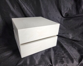 May include: A two-piece, square, white box with a subtle, swirling pattern. The box is made of a solid material and has a clean, modern design. The top half of the box is slightly ajar, revealing the interior.