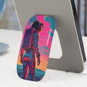 May include: A smartphone with a pop-up phone grip featuring a colorful illustration of an astronaut. The astronaut is wearing a spacesuit and standing against a vibrant sunset backdrop. The phone is gray and is propped up on a white surface.