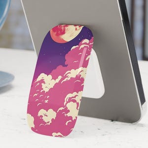 May include: A mobile phone is supported by a phone grip with a vibrant design. The grip displays a pink and purple sky with a full moon and clouds. The phone is grey and the grip is white when in use.