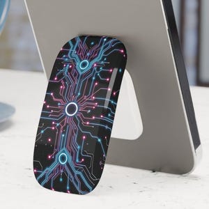May include: A smartphone with a black and white circuit board phone grip. The grip features a design of interconnected lines and circles in blue and pink. The phone is resting on a white surface, with a blue mug in the background.