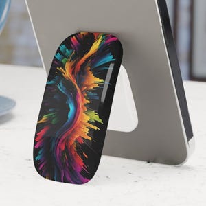 Puede incluir: Un smartphone plateado con un agarre para teléfono de diseño abstracto colorido, apoyado sobre una superficie blanca. El agarre del teléfono presenta un vibrante remolino de rojo, amarillo, azul y verde sobre un fondo negro. Una taza azul está en el fondo.