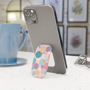Puede incluir: Un smartphone gris con un soporte para teléfono desplegable colorido con un patrón de círculos de colores pastel. El teléfono está apoyado sobre una superficie blanca, con una taza azul y obras de arte enmarcadas en el fondo.