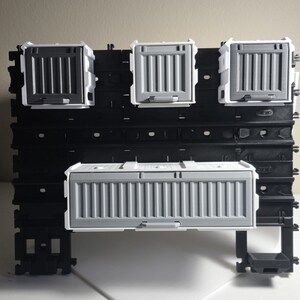 May include: A black pegboard with four gray and white miniature shipping container models attached. The containers have ribbed sides and are mounted on the board. The pegboard is on a white surface.